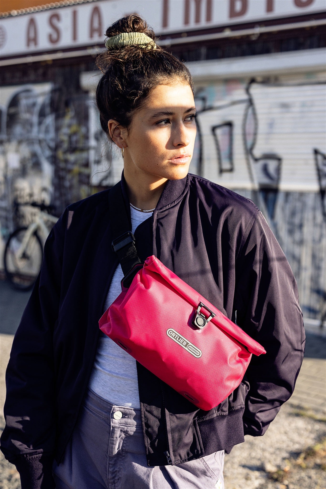 Porträt einer Frau mit ORTLIEB Velo-Sling Flex Umhängetasche in cyber-pink vor urbaner Kulisse.